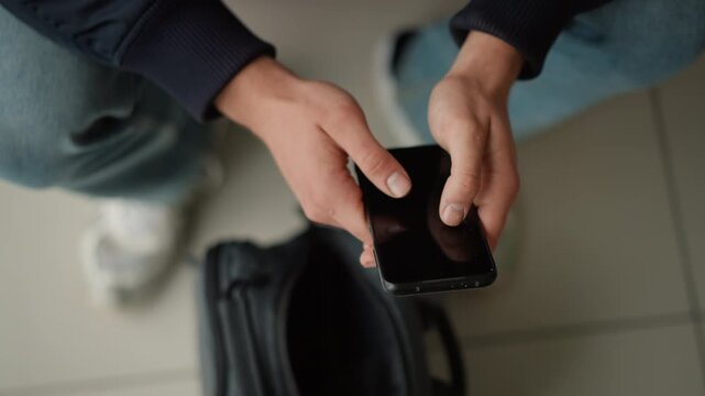 Close up of person hands holding and interacting with smartphone over open black bag on floor. person wears blue jeans white shoes and part of their dark colored jacket is visible. Soft