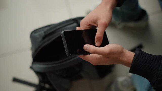 Close up of person holding and interacting with smartphone over open black backpack on light colored floor. person wearing dark clothing manipulates device while seated or standing
