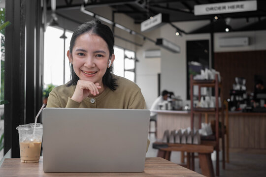 Asian woman smiling at the camera while working on a laptop in a modern cafe, with wireless earbuds and coffee, representing remote work, digital lifestyle, productivity, technology, and business.