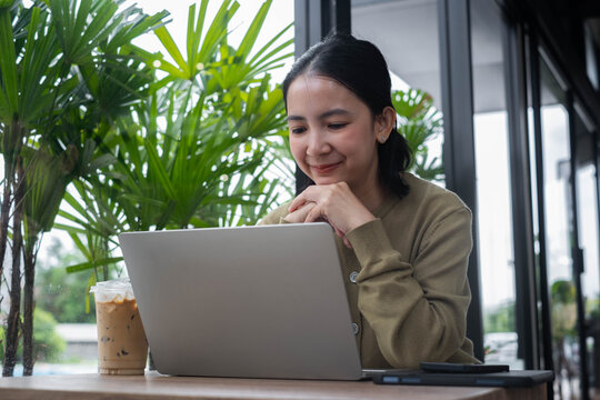 Asian woman working on a laptop in a modern cafe with coffee, enjoying a productive moment. Lifestyle scene representing remote work, freelance business, technology, productivity, work-life balance.
