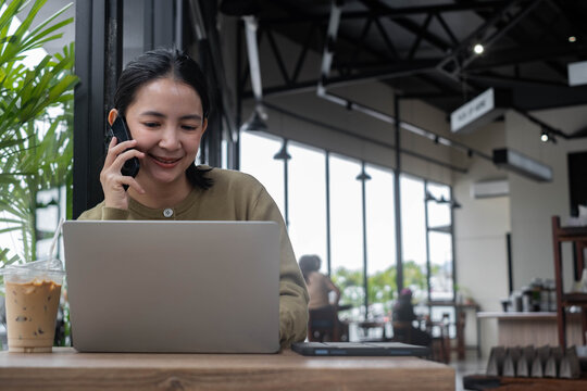Asian woman working on a laptop while talking on a smartphone in a modern cafe, enjoying coffee during a productive work. Concept of remote work, business, communication, technology, flexibility.