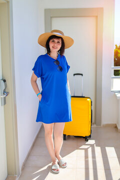 Happy woman tourist in blue dress and sun hat with bright yellow suitcase at sunny hotel near her room enjoying summer vacation travel.
