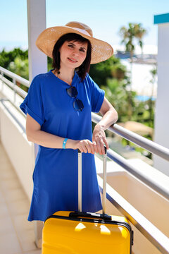 Happy woman tourist in blue dress sun hat with bright yellow suitcase at sunny hotel near her room enjoying summer vacation and travel.