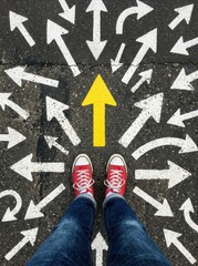 Fototapeta premium Person in Red Sneakers Standing on Asphalt with Many White Directional Arrows and One Yellow Arrow Pointing Forward