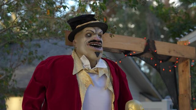 Close-up of a grinning Halloween ringmaster prop in a red coat and top hat by a suburban porch. Orange string lights and a gold skull create a spooky outdoor decoration.