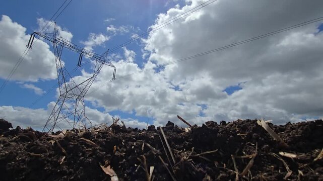 Plowed field texture of dark compost soil below a towering high voltage pylon grid system under a fast moving blue sky time lapse, capturing industrial integration with rural earth.