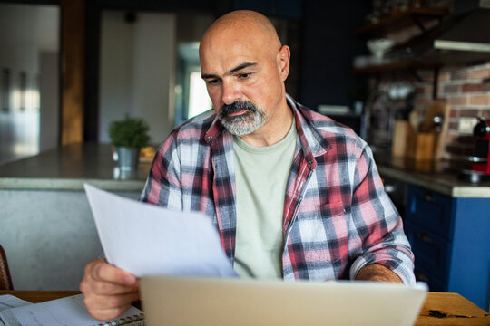 Middle-aged man reviewing household bills on laptop in home kitchen