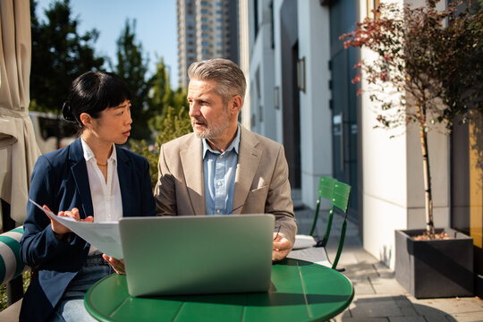 Two business professionals reviewing documents with laptop at outdoor cafe