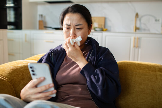Sick woman with tissue checking smartphone on sofa at home