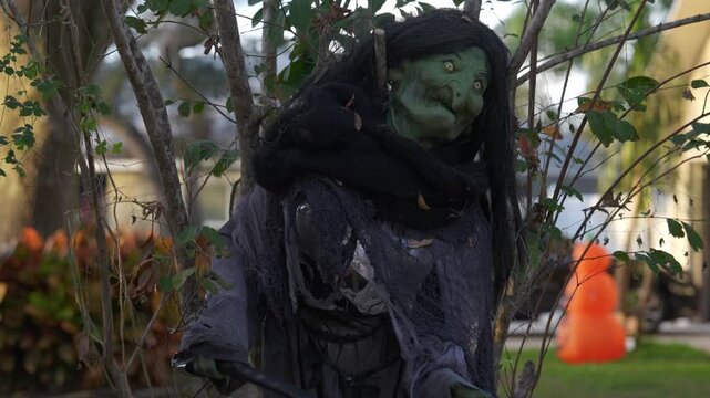 Spooky green-faced witch prop with long black hair and tattered gray robes stands among branches holding a broom. Halloween yard decoration in evening light creates a creepy haunted scene.