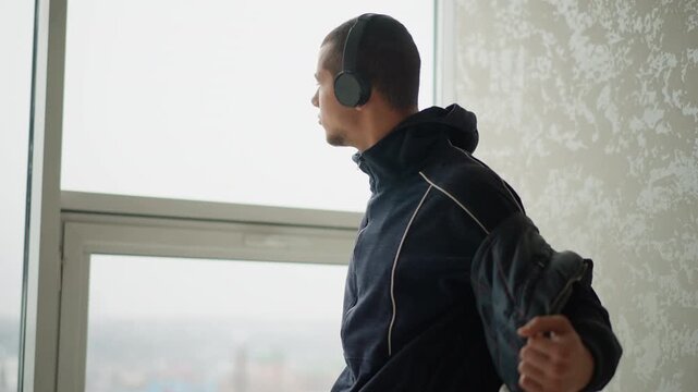 Man wearing headphones and dark jacket stands by window gradually removing his outer layer to reveal inner hoodie. scene features soft natural light from large glass window with textured