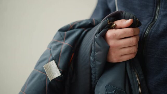 Close up of person holding and adjusting collar of blue quilted jacket with orange stitching wearing dark zip up hoodie. hand grips fabric near metal zipper pulls while another arm rests