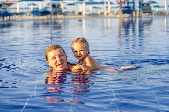 Happy mother and young daughter playing together in blue swimming pool during sunny summer vacation at tropical resort, enjoying quality family bonding time in water.