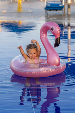 Adorable little girl having fun in swimming pool while riding bright pink inflatable flamingo pool float during sunny summer vacation day with clear blue water