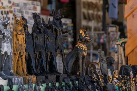 Collection of ancient Egyptian themed statues including figures with animal heads like Anubis and Bastet, pharaohs, and other deities, displayed for sale in market setting with shallow depth of field