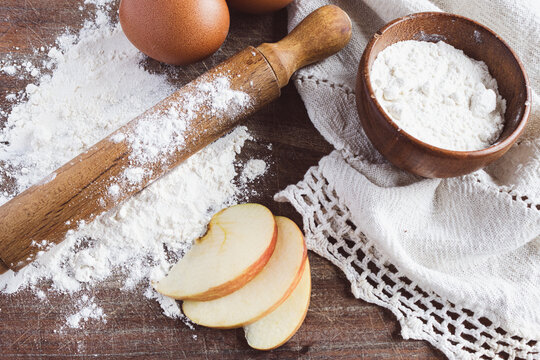 A selection of delicious ingredients for gourmet cakes and desserts, including flour, apple slices, eggs and a rolling pin, all presented on a rustic wooden board.