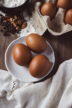 Fresh eggs and various baking ingredients like flour, chocolate chips, and nuts are arranged on a rustic wooden surface, preparing for a homemade cooking or baking project