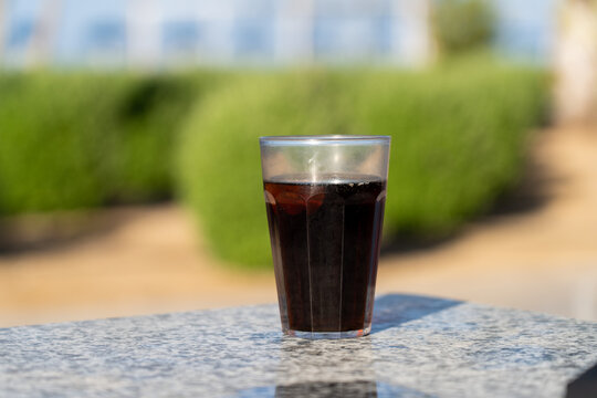 Refreshing glass of dark carbonated soda beverage sitting on speckled outdoor stone table against soft green garden background on sunny day with natural light
