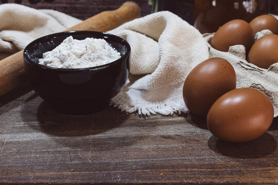 Baking ingredients on rustic wooden table preparing dough, flour in bowl with rolling pin and fresh organic eggs in carton for homemade cooking recipe