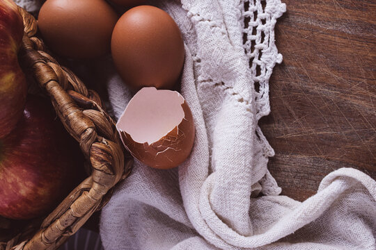 Fresh organic ingredients for home baking and healthy cooking, featuring whole apples, brown eggs, and a cracked eggshell in a woven basket on a rustic wooden table