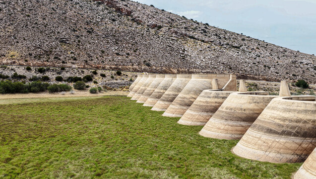 Aerial view of the concrete buttress dam wall in the Little Karoo, surrounded by green fields and rocky hills in Garden Route District Municipality, Western Cape, South Africa.