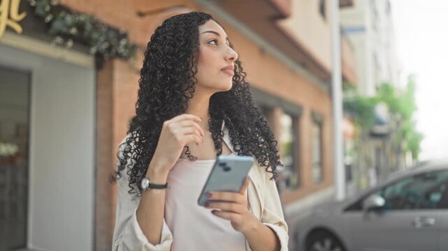 Woman holding and tapping smartphone in hands, looking up on street outside a storefront and parked car; thoughtful curiosity.