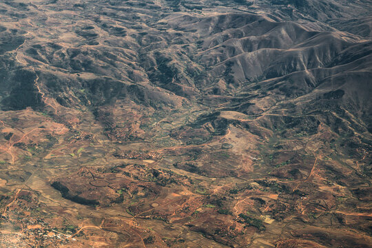 Aerial view of the rugged, mountainous landscape featuring rolling hills, terraced fields, and winding dirt roads visible from a flight over the central highlands of Madagascar.
