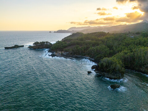 Aerial view of the rocky coastline with lush green vegetation and waves crashing against cliffs during golden hour in Pacitan, East Java, Indonesia.