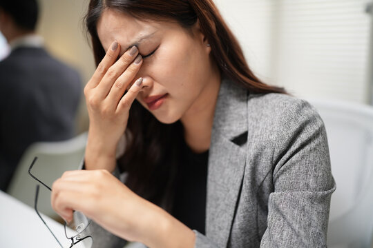 Businesswoman holding glasses and rubbing forehead experiencing work-related stress and eye strain fatigue in office environment