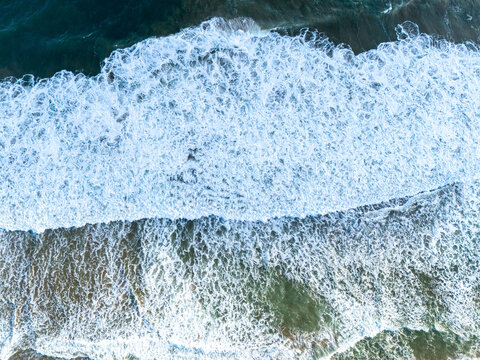 Aerial view of ocean waves crashing onto the sandy shore with white sea foam and turquoise water in Pacitan, East Java, Indonesia.