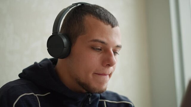 Close up of young man wearing black over ear headphones and dark blue hoodie focused on screen with slight head movement. Soft lighting highlights calm expression against light colored wall