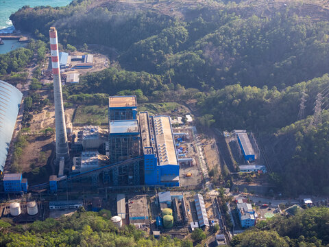 Aerial view of a power plant with a tall chimney and industrial buildings nestled among lush green hills near the coast in Pacitan, East Java, Indonesia.