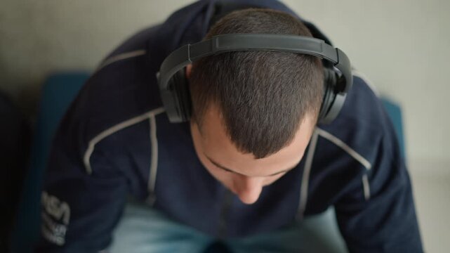 Close up of man wearing headphones and looking down with his head bent forward. subject is dressed in dark clothing seated on blue furniture against indoor background. Soft lighting creates