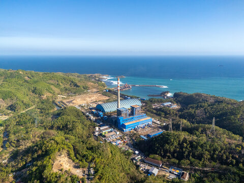 Aerial view of the coal-fired power plant situated along the coastline, featuring a tall chimney, industrial buildings, and surrounding lush green hills in Pacitan, East Java, Indonesia.