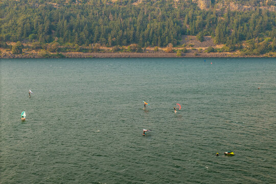 Aerial view of windsurfers and wingfoilers gliding across the Columbia River under a hazy sky with forested hills in the background in Hood River, Oregon, United States.