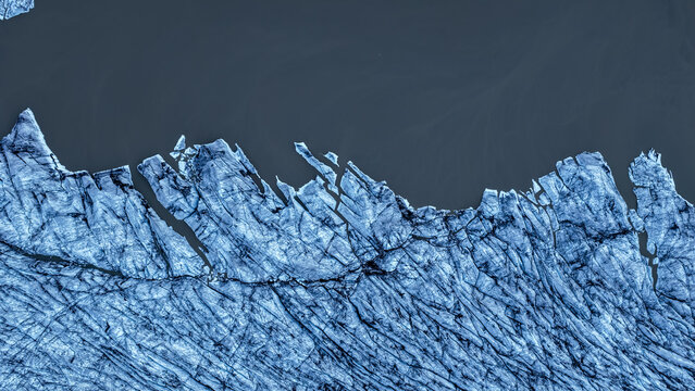 Aerial view of Fjallsarlon glacier with deep blue crevasses and jagged ice formations meeting the dark glacial lagoon waters Hofn, Sveitarfelagid Hornaf, Iceland.