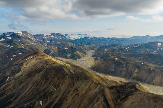 Aerial view of Fjallabak Nature Reserve with dramatic volcanic mountains, winding river valleys, and patches of snow under a cloudy sky in Rangarthing ytra, Iceland.