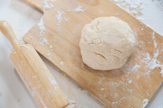Dough is prepared on a wooden board with a rolling pin nearby in a kitchen