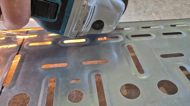 Side close-up shot of a worker cutting the edge of a metal cable tray longways with a hand angle grinder as sparks fly.