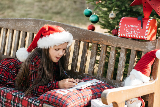 Christmas in July with girl wearing Santa hat writing letter to Santa outdoors beside decorated Christmas tree, Santa mailbox and festive decorations in sunny summer garden.