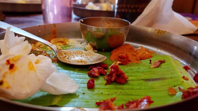 An empty steel platter with leftover scraps of a South Indian meal on a banana leaf. A used spoon, an empty small bowl, scattered food remnants, & crumpled tissues signal the satisfying end of a feast