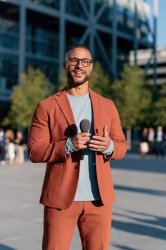 Male presenter holding a microphone in an urban plaza wearing terracotta suit for broadcast interview background and on-camera presenter banner