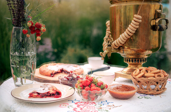 A golden samovar with bagels, tea cups, homemade pie, and jam on a white tablecloth set for tea in a garden.