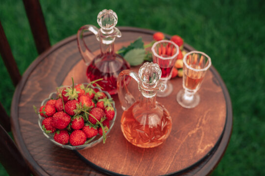 Crystal decanters with strawberry liqueur and glasses set on a wooden table, decorated with fresh strawberries in a natural garden setting.