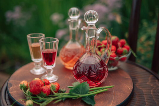 Crystal decanters with strawberry liqueur and glasses set on a wooden table, decorated with fresh strawberries in a natural garden setting.