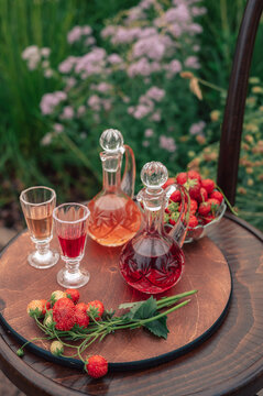 Crystal decanters with strawberry liqueur and glasses set on a wooden table, decorated with fresh strawberries in a natural garden setting.