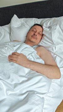 Caucasian male sleeping peacefully in bed covered with white satin sheets, resting on a dark upholstered headboard in a modern bedroom setting