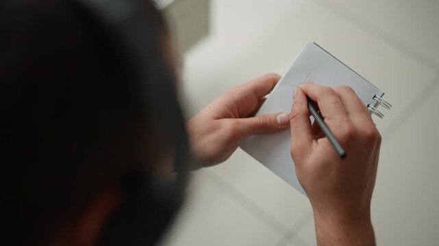 Close up of hands holding notebook and drawing with pencil on white paper. Notebook is spiral bound held steady while sketching simple line art. Bright indoor setting with soft lighting