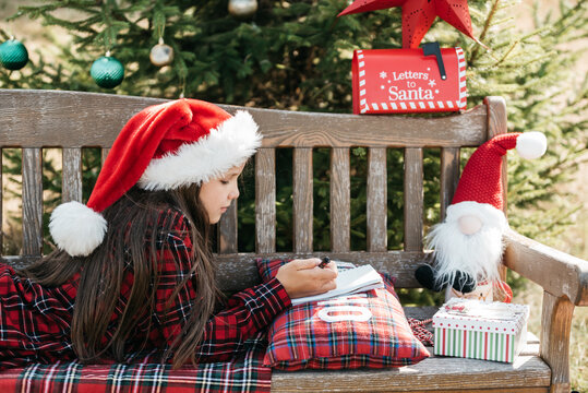 Christmas in July with girl wearing Santa hat writing letter to Santa outdoors beside decorated Christmas tree, Santa mailbox and festive decorations in sunny summer garden.