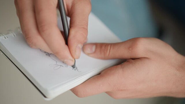 Close up of hands drawing with pencil on sketchbook page gradually adding lines to form simple sketches. Notebook held steady while artist focuses on precise hand movements and paper texture